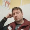 Мужчина в бордовом жилете с нашивкой Магнит на груди, сидит в светлом помещении склада, руки на лице, на запястьях черные браслеты, позади белые плитки и информационные постеры