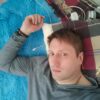 Мужчина с короткой стрижкой лежит на синем пушистом пледе, рядом тканевый клетчатый плед и белая гарнитура на подушке; спокойное выражение лица, серый свитер, цепочка на груди