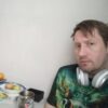 Мужчина с короткими светлыми волосами в зелёной футболке с ярким принтом и серыми наушниками вокруг шеи сидит за столом. На столе фрукты в миске, кружка с символом и мясная нарезка на тарелке. Фон нейтральная светлая стена.