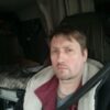 Мужчина в салоне автомобиля: бордовая куртка, светлая меховая накидка, ремень безопасности, спокойный взгляд, приглушённый свет