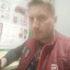 Вблизи мужчина в бордовой куртке стоит рядом с информационными стендами; позади зеленая стена, атмосфера двора и Питера