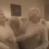 A happy elderly couple in their 60s or 70s standing together in a cozy living room, holding hands and smiling warmly at each other, wearing elegant but comfortable clothing, a beautiful wedding ring visible on the woman's finger, soft warm lighting, symbolizing a long and lasting marriage