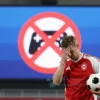 A Russian football fan in a stadium, wearing a red and white jersey, holding a football, looking disappointed at a large digital screen that displays a stylized banned video game icon (a crossed-out controller), with no text or lettering visible
