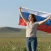 A woman standing in an open field, holding a Russian flag aloft. She is wearing casual, modest clothing, with a calm and proud expression. The background shows a wide green meadow under a clear blue sky, with gentle hills in the distance. The focus is on the woman and the flag, with natural lighting highlighting the scene.
