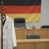 A realistic scene set in a German courtroom, focusing on a white lab coat hanging on a wooden coat rack in the foreground. In the background, a judge's bench with a gavel and subtle lighting that hints at the colors of the German flag, but without any visible text or symbols. The atmosphere is serious and investigative, with soft shadows and a photorealistic quality.
