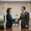 A diplomatic scene showing representatives from Russia and Mexico shaking hands over a large steel coil, with the Russian and Mexican flags subtly displayed in the background, and symbols of alternative currencies or digital tokens floating nearby, emphasizing a partnership to bypass the US dollar in steel transactions. The setting is a modern conference hall with a sleek, professional atmosphere, no visible text or logos.