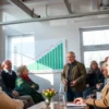 A warm community meeting in a modern office in Astrakhan, with a knowledgeable official standing beside a large transparent chart showing rising pension amounts, surrounded by attentive elderly men and women of diverse backgrounds, bright natural light through windows, realistic details, no text or signage