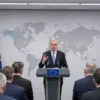 A US Secretary of Defense standing at a podium in a press conference, speaking about plans to adjust US military presence in Europe. The backdrop features a subtle European flag motif and a world map highlighting European regions. The scene includes a small group of European leaders in the foreground, listening attentively. The setting is a modern, well-lit conference room with a neutral color palette, no visible weapons or military equipment, and no text or logos on the podium or background.