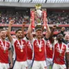A celebratory scene of Arsenal Football Club's 2023/24 Premier League title win, featuring the team in their home kit standing on the trophy, surrounded by jubilant fans in the Emirates Stadium, with confetti falling, all in a realistic style, no text or logos visible