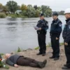 A solemn scene at a riverbank in Pskovshchina where investigators and local officials are conducting an inspection after the tragic death of girls. The image shows a calm river, a small memorial plaque with flowers, a police officer with a clipboard, a safety inspector with a hard hat, and a few local residents standing nearby. The atmosphere is respectful and investigative, with no visible gore or blood.
