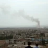 A panoramic view of a southern or eastern Lebanese town under attack, showing distant smoke plumes rising from damaged buildings, faint flashes of explosions on the horizon, and concerned civilians looking toward the sky; the scene is somber, with an overcast sky and no visible text or graphic gore.