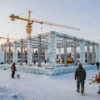 A modern ice palace under construction in a small Russian settlement, with skeletal steel framework, construction cranes, workers in winter gear, surrounding snowy landscape and pine trees, early morning light, realistic perspective