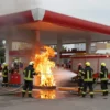 A realistic depiction of a gas station in Pyatigorsk with firefighters actively extinguishing a fire, showing flames, hoses, fire trucks, and emergency lights, set against a backdrop of the surrounding cityscape