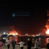 A dramatic night scene at the port of Bandar Abbas, Iran, showing large cargo ships and docked vessels with plumes of smoke and fire erupting from the water and warehouses. People in the foreground are running, covering their faces, and pointing towards the explosions, while emergency lights flash. The sky is dark with orange and red hues from the blasts, and the water reflects the flames. The atmosphere is chaotic and tense, capturing the panic and urgency of the moment.