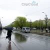 A realistic depiction of Saransk city on a rainy day in late May, with gentle drizzle, overcast sky, wet streets reflecting the surroundings, people walking with umbrellas, light green trees beginning to bud, and a subtle sense of a mild 14°C temperature, without any visible text or signage.