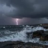 A dramatic coastal scene of the Pomorje region under a stormy sky, with dark, heavy clouds, occasional bright lightning strikes illuminating the sea and rugged shoreline, waves crashing against rocks, and a moody, atmospheric color palette