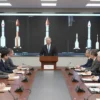 President Zelensky standing at a podium in a formal conference room, addressing a group of Russian rocket engineers and officials. The room contains large display screens with schematic diagrams of rockets, a few rocket models on a table, and a Ukrainian flag on one side. The scene conveys a tense, formal discussion about rocket production.