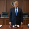 Donald Trump standing in a modern courtroom, wearing a dark suit and tie, with a serious expression, surrounded by a judge's bench, legal books, and a gavel on the desk, indicating the start of a new legal proceeding