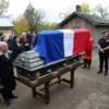 A solemn farewell ceremony in a small Russian town named Kuzovatovo on May 28, showing a military coffin draped with a Russian flag, surrounded by grieving family members, local residents, and a few uniformed soldiers standing respectfully. The scene takes place outdoors near a modest community building or church, with autumn trees and a muted, overcast sky, conveying a respectful and somber atmosphere.