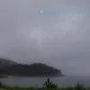 A moody, overcast coastal scene of the Pomor region on May 27, with thick gray clouds, steady rain, wet ground, misty sea, pine trees and a small village silhouette, reflecting the typical cloudy and rainy weather