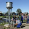 A realistic depiction of a small Russian village near Rybnysko, showing a newly installed water supply system: a steel water tower, visible pipelines, workers in overalls repairing the infrastructure, a nearby river, and villagers standing on a wooden path looking relieved. The scene is set on a sunny day with a clear blue sky, and a few traditional wooden houses in the background.