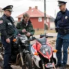 A realistic scene set in the Volgograd region showing a police officer in uniform confiscating a motorcycle from a visibly intoxicated driver standing nearby. The background includes typical Volgograd architecture or a hint of the Volga River shoreline, with a cloudy sky. The driver looks disheveled, holding a bottle loosely, while the officer secures the bike with a chain. No text or signage is visible.