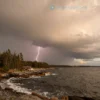 A scenic view of the Pomor region showing a warm, bright sky contrasted with dark thunderstorm clouds gathering over the sea. The landscape includes a rugged coastline, gentle waves, pine forests, and distant hills. Lightning flashes illuminate the clouds, while the overall atmosphere feels warm and dramatic, capturing the unique weather of warm thunderstorms in Pomorye.