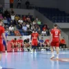 A dynamic indoor handball match scene in a modern arena in Krasnodar, showing the Lokomotiv Kuban team looking dejected after a loss, with players in motion, a tense atmosphere, spectators in the stands reacting emotionally, and the referee signaling the end of the game, all captured in vivid detail