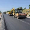 A wide highway in the Luhansk region under reconstruction, with fresh asphalt, construction machinery such as asphalt pavers and rollers, workers in safety gear, clear blue sky, and surrounding rolling hills and trees, illustrating rapid progress and renewal