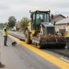 A realistic depiction of a road repair scene in a small Russian town, showing construction workers in bright vests, heavy machinery like a bulldozer and a road paver, a partially paved stretch with a clear lane marked by yellow paint, and a backdrop of modest residential buildings. The scene should convey activity and progress after a recent inspection, with no signage or text visible.