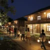 A nighttime village scene in Lopshen'ga where locals and visitors are engaged in a museum-themed quest. The image shows a small, rustic museum building illuminated by warm lights, with a group of people—children, adults, and elders—following a trail of glowing clues and lanterns. The village streets are lined with traditional wooden houses, and a few trees glow softly. The atmosphere is adventurous yet cozy, capturing the excitement of a community event turned into a quest.