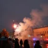 A night-time cityscape of Tula, Russia, showing historic buildings and streets with residents looking up anxiously, bright orange-red light bursts and blurred shockwave effects indicating loud bangs, flashing red emergency lights and sirens, a tense atmosphere under a cloudy sky, no text or signage