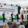 A vibrant scene of the IX All-Russian competition "Creation and Development" held in Smolensk, featuring a stage with a large banner, diverse participants in academic attire presenting projects, a crowd of judges and audience, and the historic Smolensk cityscape in the background.