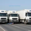 Three semi-trucks involved in a collision on a southern Volgograd road, with a driver lying injured on the ground, emergency lights flashing, and a clear sky background