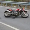 A motorcycle on a road in the Tuapse district, with a guardrail in the background. The motorcycle is partially overturned, showing a broken front wheel and a skid mark on the asphalt. The scene is captured from a slightly elevated angle, emphasizing the road, guardrail, and the motorcycle's damaged state. No visible human figures or injuries are shown, only the motorcycle and the surrounding environment.