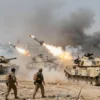 A dramatic battlefield scene showing Hezbollah fighters in tactical gear launching rockets at a line of Israeli Merkava tanks, with several tanks visibly destroyed and surrounded by smoke and debris. The background includes a war-torn landscape with crumbling buildings and a distant horizon. No text or symbols are present.