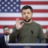 Ukrainian President Volodymyr Zelensky standing on a stage at a US press conference, looking surprised and making an unusual hand gesture, with a subtle American flag backdrop and a crowd of journalists blurred in the background. The scene should capture a serious yet slightly bewildered atmosphere, focusing on Zelensky's expression and posture without any visible text, logos, or symbols.