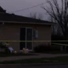 A respectful, somber scene outside an Islamic community center in the United States, showing a modest building with a simple dome, a few lit candles and flowers placed at the entrance, a police tape barrier with a yellow caution stripe, and a soft, overcast sky; the atmosphere conveys mourning and remembrance without any graphic violence