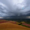 A dramatic weather scene over the Volgograd region on May 27, showing rolling fields and distant riverbanks under a dark, heavy storm cloud sky. Intense rain is falling, with visible lightning bolts streaking across the clouds and small hailstones visible in the air. The perspective is wide and horizontal, capturing the expansive landscape and the ominous atmosphere of the forecasted showers, thunderstorms, and hail.