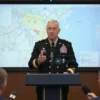 A senior military commander in a formal uniform stands at a podium in a briefing room, addressing a group of reporters and analysts. The commander is mid‑speech, gesturing with one hand while holding a microphone. Behind him, a large screen displays a map with a highlighted region, and a flag of the United States is visible in the background. The setting is professional and serious, with a focus on the commander’s expression and posture.