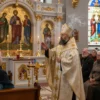 A solemn Orthodox hieromonk in traditional monastic robes stands in a richly decorated church interior, gesturing gently as he explains the significance of the Holy Trinity. Behind him, an iconic icon of the Holy Trinity (the three angels) hangs on the iconostasis, surrounded by golden candlesticks, incense smoke, and soft candlelight. Attentive parishioners, both men and women in modest attire, sit on wooden benches, listening respectfully. The atmosphere is reverent and serene, with warm, natural lighting filtering through stained glass windows depicting biblical scenes.