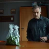 A realistic scene of a middle-aged man in his 40s standing in a modest Mordovian apartment, looking concerned, with a discreet bag of green cannabis leaves placed on a wooden table, soft indoor lighting, no text or symbols