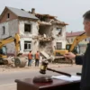 A realistic scene of a small Russian town court ordering the demolition of an illegally built house, showing a judge's gavel beside a demolition crew with excavators and wrecking balls tearing down the structure, dust and rubble on the ground, the surrounding streets of Kizlyar, without any visible text or signage