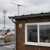 A realistic scene depicting a suburban area in Chuvashia during a maintenance-related TV and radio outage: a rooftop TV antenna, a nearby radio transmission tower, a home interior visible through an open window showing a turned-off TV with faint static, a tabletop radio with no signal, cloudy sky, subtle warning symbols like a caution triangle without text, and a calm atmosphere.