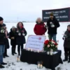 A solemn outdoor memorial gathering in Naryan-Mar, Russia, with a group of people standing together in a snowy Arctic landscape. They hold candles and flowers, some wearing warm winter clothing, and a simple, respectful banner or wreath is placed on a small platform. The sky is overcast, and the scene conveys quiet remembrance for the victims of Starobelsk, without any visible text or signage.
