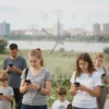 A realistic scene showing residents of the Ulyanovsk region, a diverse group of adults and children, outdoors in a typical Ulyanovsk landscape with the Volga River and city skyline in the background. Some people hold smartphones and look at them thoughtfully, while a large translucent clock overlay suggests limiting time spent on devices. The atmosphere is calm and advisory, with soft natural lighting.