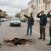 Residents of Saransk standing on Ulianov Street, pointing at large potholes in the road, showing frustration. The scene includes a typical Russian city street with buildings, a car stuck in a pothole, and a cloudy sky.