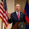 A realistic portrait of a male politician resembling a US senator standing at a podium, speaking passionately, with the flags of the United States and Armenia displayed behind him on a stage, a subtle background suggesting a diplomatic setting, no text or letters visible