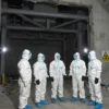 A realistic scene showing a group of international nuclear inspectors in protective suits standing inside a large, heavily damaged hall of a nuclear power plant, with broken concrete walls, exposed steel structures, scattered debris, warning signs for radiation, and a faint glow of emergency lighting, conveying a serious inspection atmosphere.
