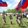 A lively outdoor scene of children of Federal Penitentiary Service employees in the Pomor region participating in a Zarnitsa competition: kids in simple scout-like uniforms running, climbing ropes, crawling under nets, and aiming mock airsoft rifles at distant targets, with bright flags fluttering, a clear sky, and a cheering crowd of families in the background. The atmosphere is energetic and patriotic, emphasizing teamwork and youthful enthusiasm.